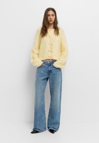 Sárga, bolyhos, rövid kardigán gombos záródással, amelyet világoskék, bőszárú farmerral párosítottak, finom hímzett mintákkal. Fekete, hegyes orrú cipők.