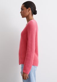 Roze gebreide trui met lange mouwen, een ronde hals en een gestructureerd patroon. De zoom is iets langer aan de achterkant, gecombineerd met lichtblauwe jeans.