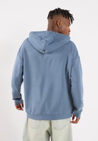 Felpa blu con una consistenza morbida, dotata di cappuccio con coulisse e polsini a costine. Indossata con jeans chiari a vestibilità comoda.
