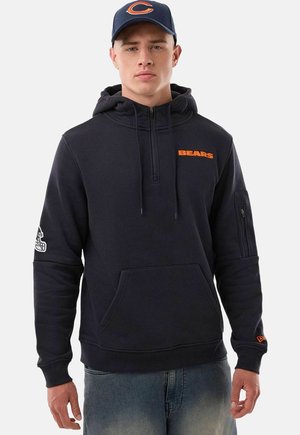 Giovane uomo che indossa una felpa nera Bears con cappuccio e un cappellino da baseball blu navy con logo arancione, in piedi davanti a uno sfondo chiaro e uniforme.