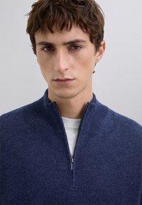 Jonge man met kort bruin haar, gekleed in een marineblauwe kwart-ritssweater over een lichtgrijze shirt, neutrale achtergrond.