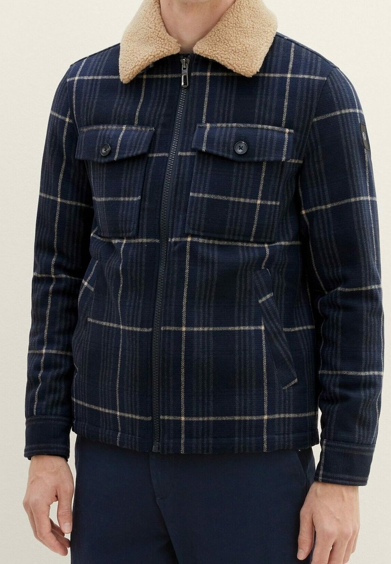 Veste à carreaux bleu marine avec col sherpa beige, fermeture éclair devant, poches poitrine boutonnées et poches sur les coutures latérales, portée par une personne debout.