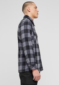 Sivá a čierna károvan á flanelová košeľa s golierom, dlhými rukávmi, zapínacím vreckom a voľným strihom. Materiál pôsobí mäkko a má textúru.