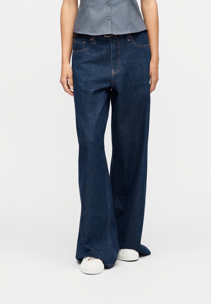 Femme portant un jean denim bleu foncé à jambes larges, une chemise grise courte boutonnée et des baskets blanches, debout contre un fond uni.