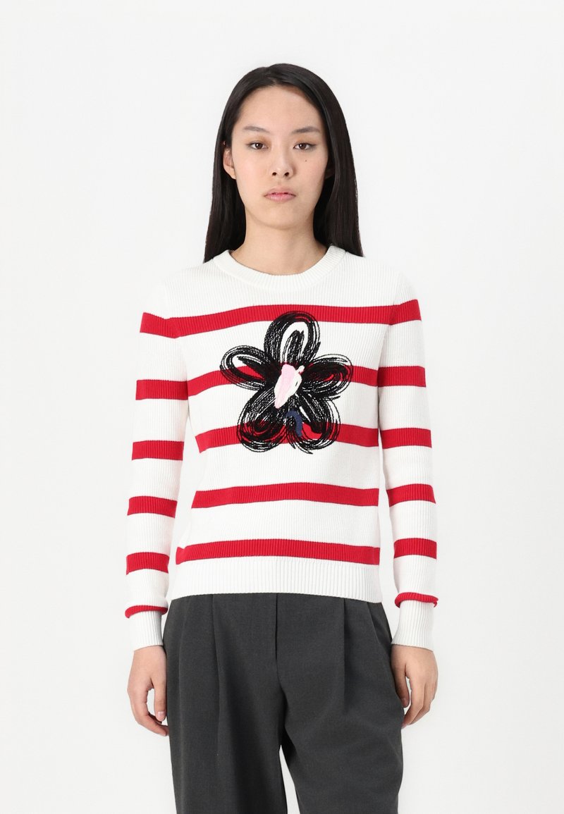 Frau trägt einen weißen Pullover mit roten horizontalen Streifen und einem großen schwarzen und pinken Blumenmuster in der Mitte, kombiniert mit dunkelgrauen Hosen.