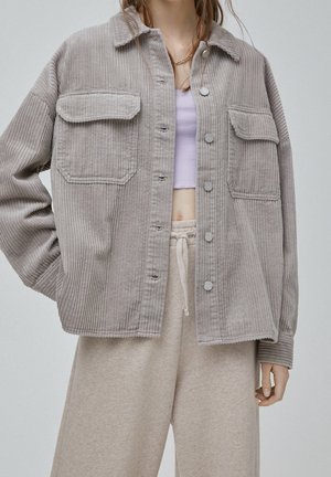 Woman wearing light gray corduroy jacket with button closure, beige drawstring pants, and a light purple cropped top.