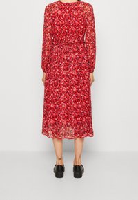 Robe midi rouge à motifs floraux avec manches longues, taille froncée et tissu transparent. Chaussures plateformes noires complètent la tenue.