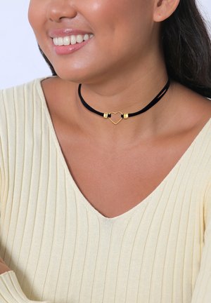 Schwarzes Samt-Choker mit einem goldenen Herz-Anhänger und zwei metallischen Akzenten, getragen über einem hellgelben gerippten Pullover.