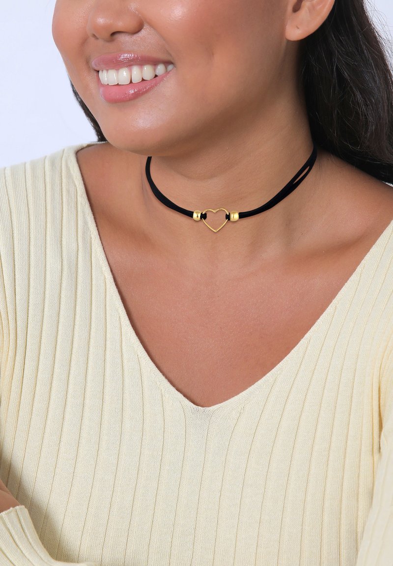 Zwarte fluwelen chokersluiting met een gouden hartbedel en twee metalen accenten, gedragen over een lichtgele geribde trui.