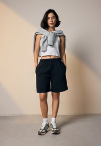 Schwarze Baumwollshorts mit elastischem Bund, grauer Pullover um die Schultern gelegt und grau-weiße Turnschuhe mit braunen Akzenten.