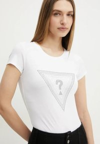 T-shirt blanc à manches courtes avec un motif triangulaire à pois et un point d'interrogation au centre. Le tissu semble doux et léger.