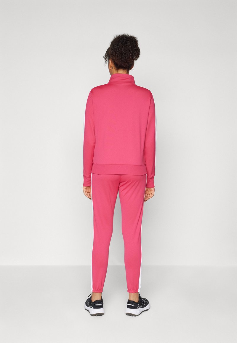 Tuta sportiva rosa con colletto alto, strisce bianche sui pantaloni e polsini elastici. Il modello è in piedi con la schiena rivolta verso l'osservatore.