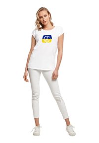 Weißes T-Shirt mit aufgerollten Ärmeln, das ein blaues und gelbes Frieden-Symbol-Graphic zeigt. Das Model trägt eng anliegende weiße Hose und weiße Sneakers.