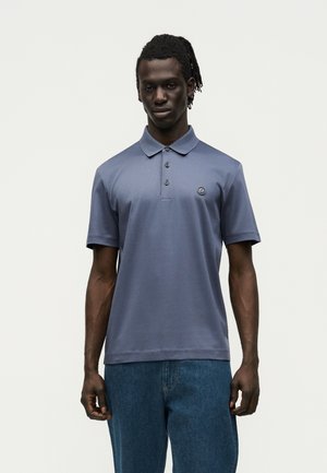 Man with dreadlocks wearing a blue short-sleeve polo shirt and blue jeans, standing against a plain light background.