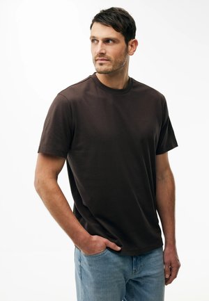 Hombre con camiseta de manga corta de color marrón oscuro y jeans azules, con la mano izquierda en el bolsillo, mirando hacia su derecha sobre un fondo blanco.