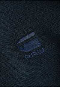 Tela azul marino con una textura tejida, que presenta un logo bordado en un tono ligeramente más claro que dice "RAW". Detalle en primer plano.