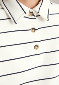 Witte polo met dunne horizontale zwarte strepen en drie bruine knopen op de sluiting onder een klassieke kraag.