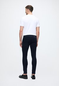 Tmavomodré úzke džínsy s hladkou textúrou, s klasickým dizajnom piatich vreciek a minimálnymi stehmi, nosené s jednoduchým bielym tričkom.