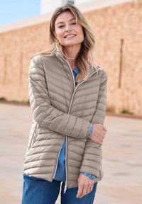 Blonde vrouw glimlachend, gekleed in een lichtgrijze gewatteerde jas over een spijkerhemd en blauwe jeans, staand in de buitenlucht bij een beige muur.