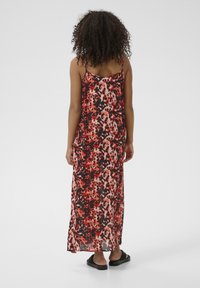 Robe maxi avec un motif léopard vibrant en rouge, noir et rose, des bretelles fines, et un tissu léger et fluide. Associée à des sandales noires.