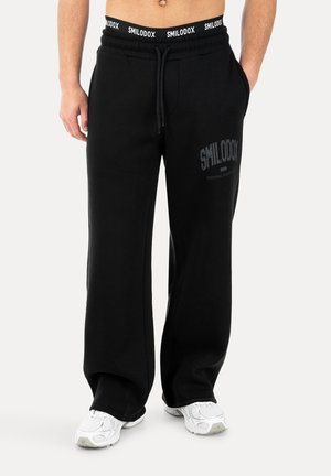 Mann trägt schwarze locker sitzende Smilodox Sweathosen mit Logo am Oberschenkel und Bund, kombiniert mit weißen Sportschuhen, Hände in den Taschen.