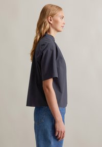 Maglietta grigia a maniche corte con colletto alto, realizzata in tessuto leggero, caratterizzata da una vestibilità comoda e una texture liscia. Abbinata a jeans blu.