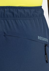 Dunkelblaue Sportshorts mit elastischem Bund, ausgestattet mit einer kleinen Reißverschlusstasche und dem "Rossignol"-Logo in Hellgrau an der Seite.