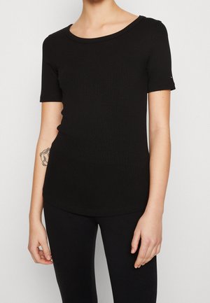 Femme portant un haut noir côtelé moulant à manches courtes et un pantalon noir, montrant un tatouage sur l'avant-bras gauche sur un fond uni.