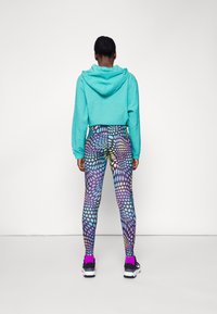 Hoodie cropped turchese in tessuto testurizzato, abbinato a leggings colorati con motivo a pois in viola, rosa e blu. Scarpe da ginnastica nere.