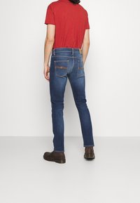 Jeans denim biru tua dengan potongan ramping, dilengkapi saku belakang dan jahitan coklat muda yang halus. Dipadukan dengan kemeja lengan pendek merah dan sepatu boot coklat.