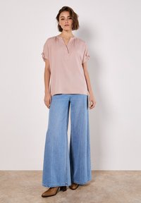 Helles rosa Bluse mit V-Ausschnitt und kurzen Ärmeln, die mit Rüschendetails verziert ist, kombiniert mit weit geschnittenen, hellblauen Jeanshosen und dunkelbraunen Stiefeln.