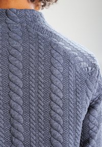 Suéter gris de punto con patrones de cable elevados. Acabado texturizado con hilos entrelazados, que presenta un escote estilo crew y detalles sutiles.