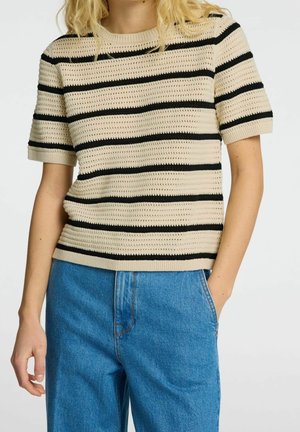 Woman wearing beige crochet short-sleeve top with black horizontal stripes and blue high-waisted denim jeans, hand in pocket.
