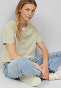 T-shirt beige a maniche corte con scollo rotondo, abbinato a jeans azzurri chiari. Il tessuto appare morbido, con una vestibilità comoda.