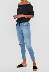 Top negro sin hombros con detalle de botones y cuello elástico, combinado con jeans skinny azul claro con roto en la rodilla y sandalias negras tipo slide.