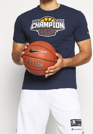 Hombre sosteniendo un balón de baloncesto Nike Elite, vistiendo una camiseta Champion azul marino con el logo de baloncesto y pantalones cortos blancos Champion con parche de logo.