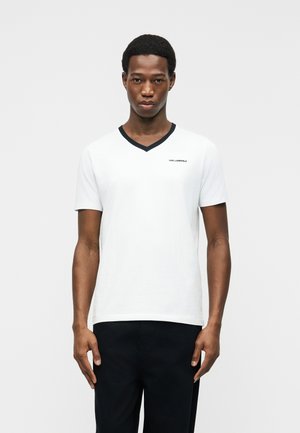 Homme portant un T-shirt blanc à col en V avec bordure noire et petit logo "KARL LAGERFELD", associé à un pantalon noir, debout devant un fond uni.