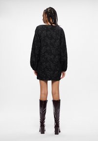 Melna kleita ar ziedu rakstu, garām piedurknēm, teksturēta auduma un brīvas piegriezuma. Valkāta ar ceļgala augstuma, spīdīgām, tumši brūnām zābakām.