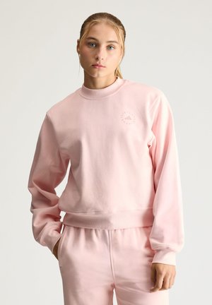 Kvinde med blondt hår i hestehale iført en lyserød sweatshirt og matchende bukser, stående foran en ensfarvet lys baggrund.