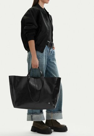 Žena v černé bundě, ohrnutých modrých džínách a botách držící velkou černou koženou kabelku za úchyty.