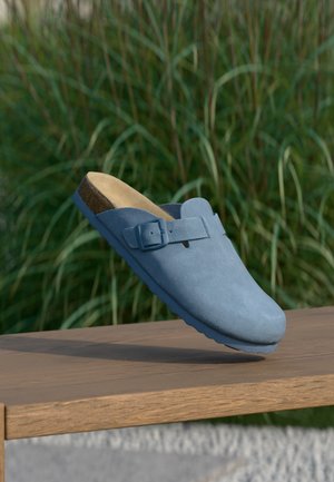 Blaue Wildleder-Clogs mit abgerundeter Spitze und seitlicher Schnallendetail, auf einer Holzoberfläche platziert, mit strukturierter Sohle und Korkeinlegesohle.