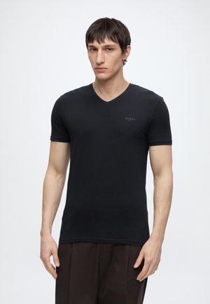Jeune homme portant un t-shirt noir moulant Guess à col en V et un pantalon brun foncé, debout devant un fond blanc uni.