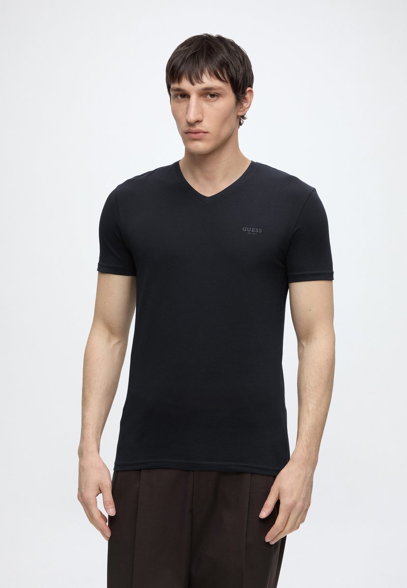 Jeune homme portant un t-shirt noir moulant Guess à col en V et un pantalon brun foncé, debout devant un fond blanc uni.