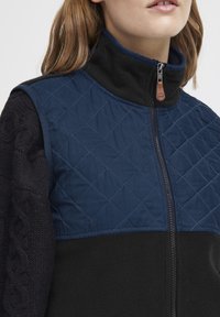 Gilet matelassé avec une partie supérieure bleu marine à motif losange et une partie inférieure en polaire noire. Fermeture éclair avec un tirant en cuir.