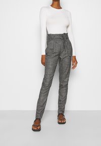 Graue kariierte hochtaillierte Hose mit einem taillierten Bund, schmal zulaufenden Beinen und einem strukturierten Stoff. Kombiniert mit einem enganliegenden weißen Langarmoberteil.
