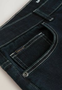 Jeans din denim închis, cu un buzunar rotund cu cusături albe, o etichetă mică etichetată "SUPPLY CO" și detalii cu un buton metalic.