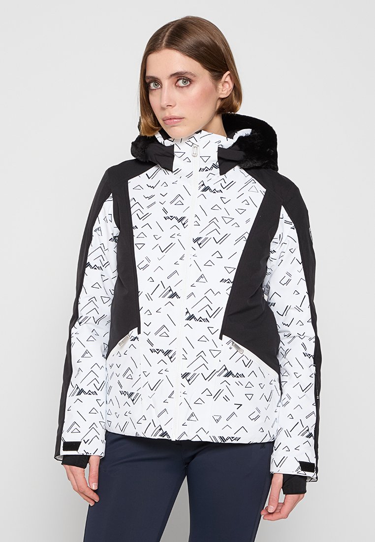 Kobieta w białej i czarnej wzorzystej zimowej kurtce z kapturem wykończonym futrem oraz granatowych spodniach, stojąca na tle jednolitym.