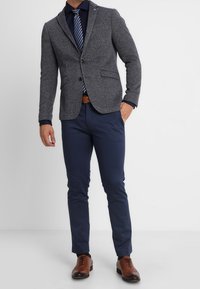 Uomo in giacca grigia, camicia blu con cravatta a righe, pantaloni blu, cintura marrone e scarpe eleganti marroni, in piedi con una mano in tasca.