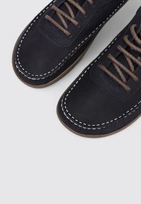Mocasines de cuero negro con cordones marrones y costuras blancas alrededor de la punta sobre un fondo gris claro.