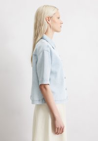 Light blue denim short-sleeve shirt with a button front, pointed collar, and a relaxed fit. Fabric appears textured with visible stitching.